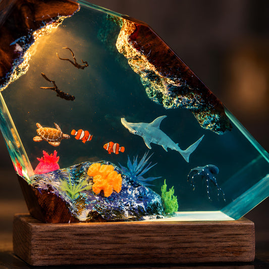 Ocean resin lamp with hammerhead shark, two scuba divers, sea turtle, clownfish, octopus, coral reef details, blue LED glow, and natural wood base.